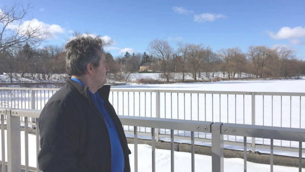 A middle age man in a black jacket with a blue fleece sweater under it, standing beside a metal fence looking out onto a snow-covered frozen pond.