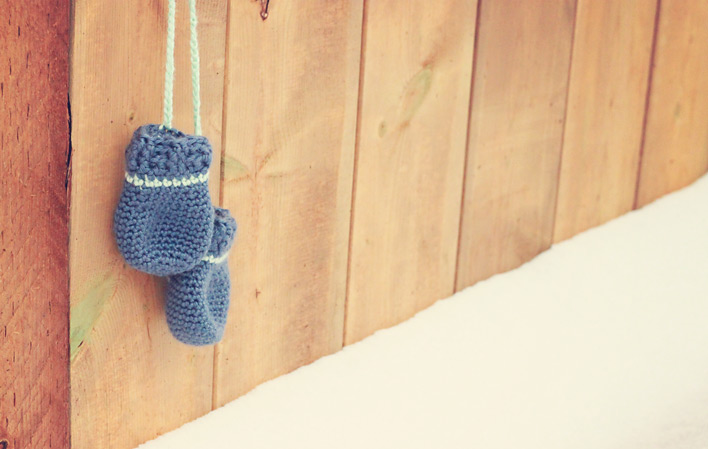 Tiny blue crocheted fingerless mittens for a newborn baby, hanging on a wooden fence by a light green string.