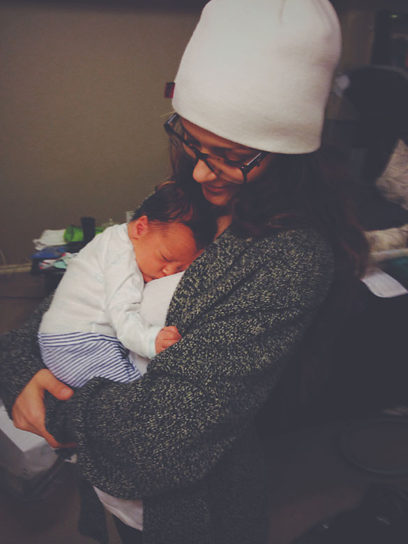 A young woman with a white toque and oversized knitted black cardigan, holding a newborn baby dressed in a white long sleeve diaper shirt and blue and white striped pants.