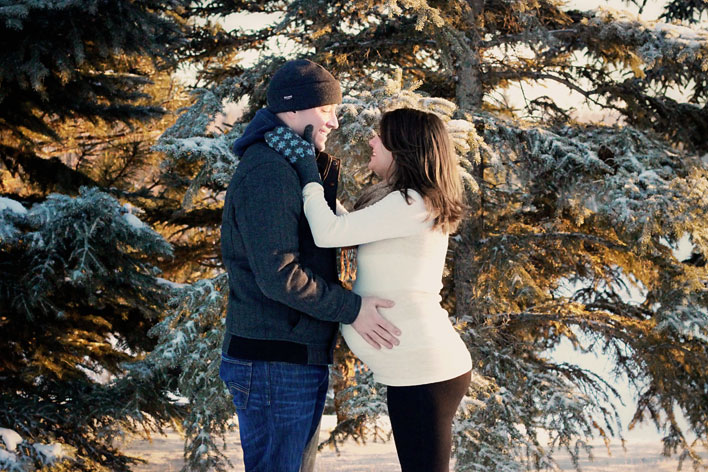 A pregnant woman standing sideways facing her husband with her gloved hand on his cheek. He is facing her, wearing a toque, and has his hand on her pregnant belly. They are standing in front of a snow-covered spruce tree.
