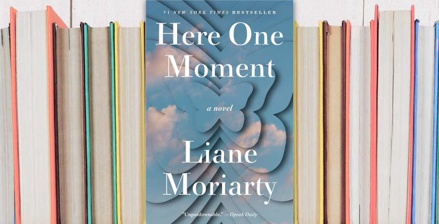 A shelf of rainbow books with the pages facing out, with Here One Moment by Liane Moriarty in the middle with the cover facing forward.