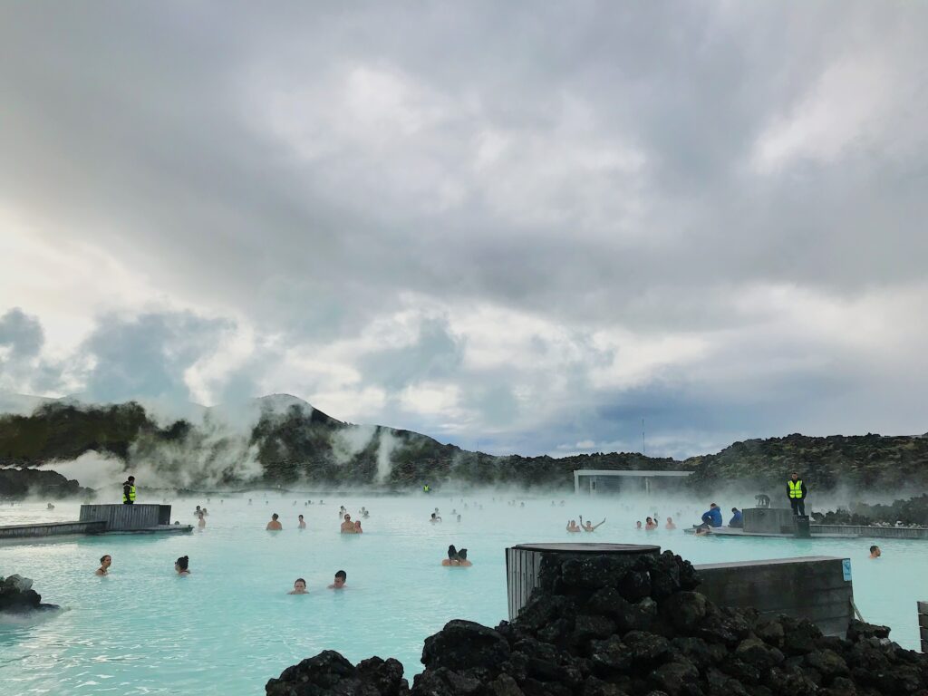 The Blue Lagoon in Iceland, a big pool of turquoise water with steam coming off of it, surrounded by black lava rock. There are about 50 people in the water.