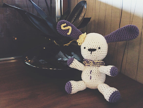 A cream coloured crocheted bunny with purple ears, with a yellow S on the left ear, and a crocheted yellow flower on its head.