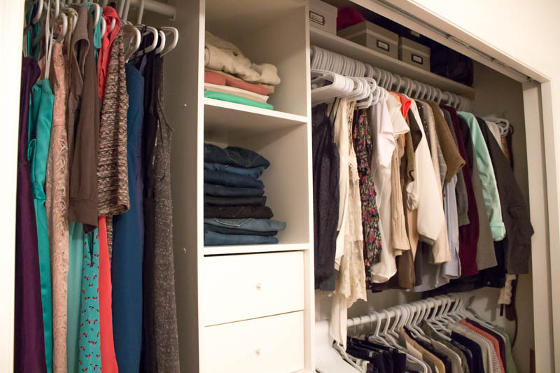 A close up of the closet organizer, with dresses, shirts and sweaters hanging on hangers, photoboxes on the top shelves, and pants folded on the shelving unit.