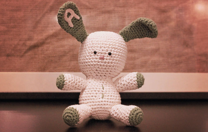 A crocheted bunny, cream in colour, with an oversized head and floppy green ears. The left ear has a capital A crocheted applique attached to it. 
