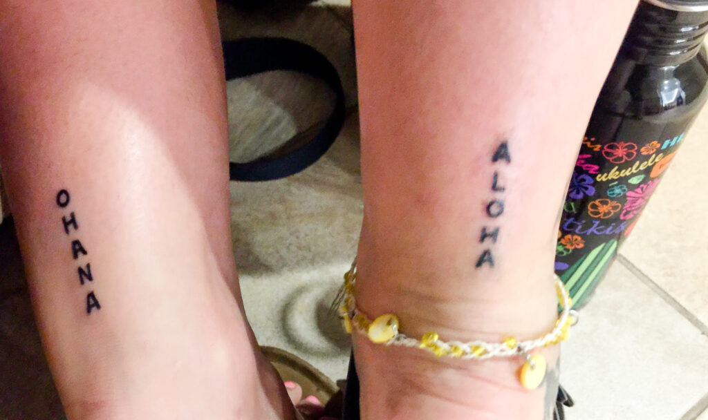 The back of two ankles with matching maui themed tattoos. The left back of the angle says 'ohana' in capital letters and black ink, the right back of the ankle says 'aloha' in capital letters. It's also wearing a yellow anklet.