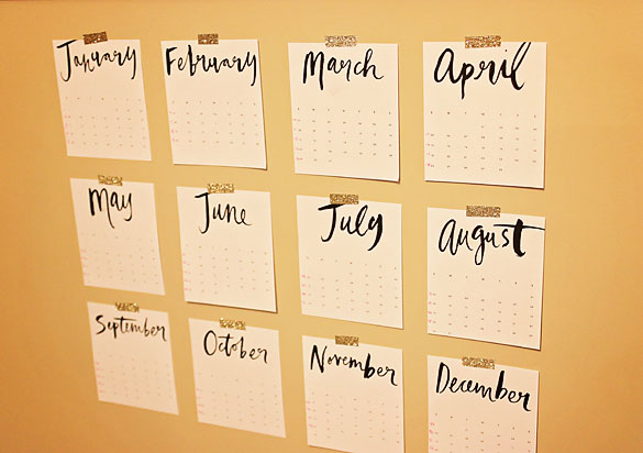 12 cut out month calendars with a script font, taped to a wall with gold glitter tape.