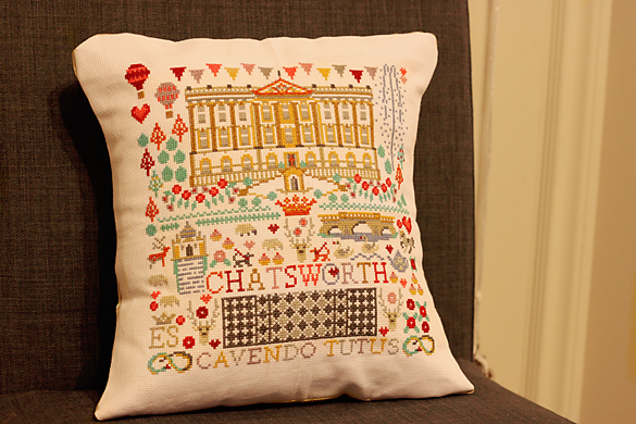 A cross-stitched pillow of different elements related to Chatsworth House in Chesterfield, England, propped up on a chair.