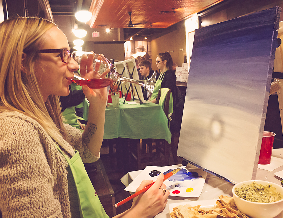 A woman with blond hair sipping a glass of rose wine while painting a canvas.