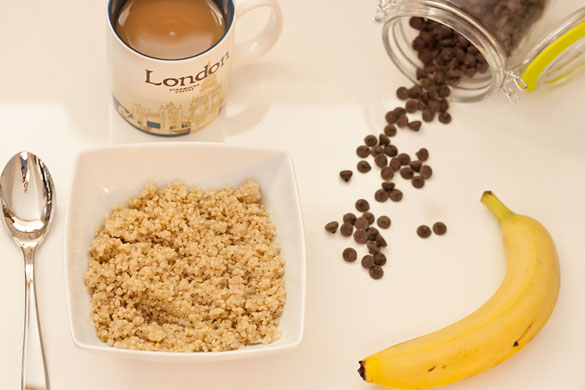A bowl of quinoa on a white glossy table with a silver spoon, a jar with chocolate chips spilling out, a cup of coffee with milk, and a banana.