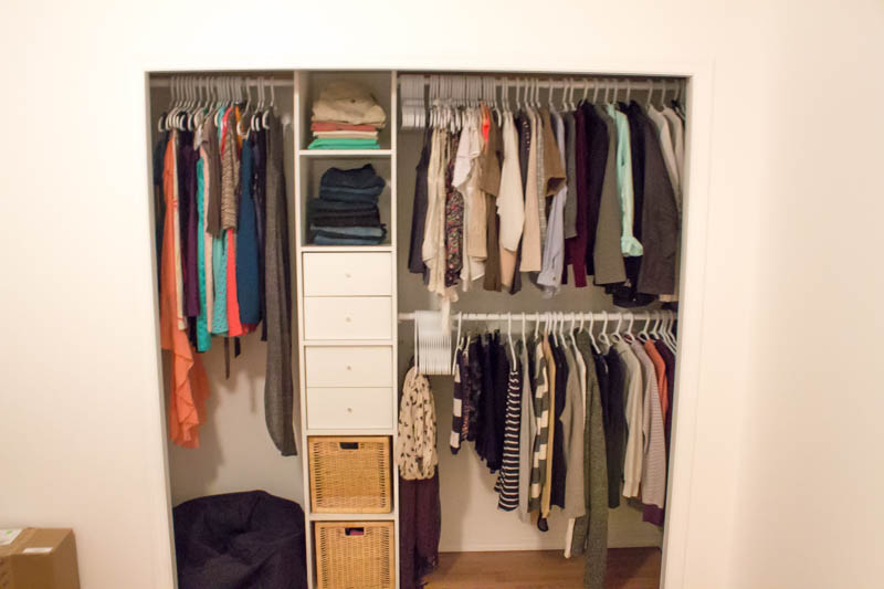A picture of a closet with a built in organizer. There's a tall hanging space on the left side, with two hanging spaces on the right side. It's split in the middle by a shelving unit with 6 shelves. The top two are open and have folded pants, the middle two have a 2-drawer insert in each, and the bottom two have a wooden basket in them. 