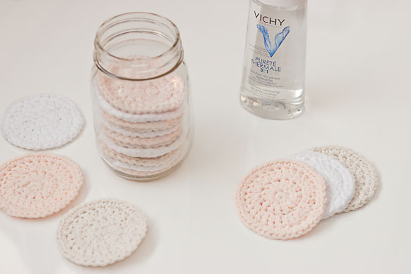 A glass mason jar filled with crocheted reusable makeup remover pads that are white, peach. and off-white in colour. There is also a bottle of micellar water on the counter. 