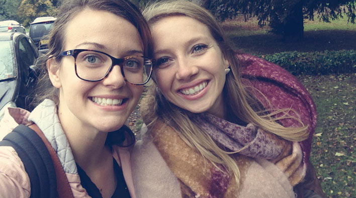 Becca and Natasha on a walk through the Countryside in England. Becca has brown hair, pulled back into a low bun and is wearing a pink jacket and glasses. Natasha has long blonde hair and a big, cozy burgandy and beige scarf around her neck.