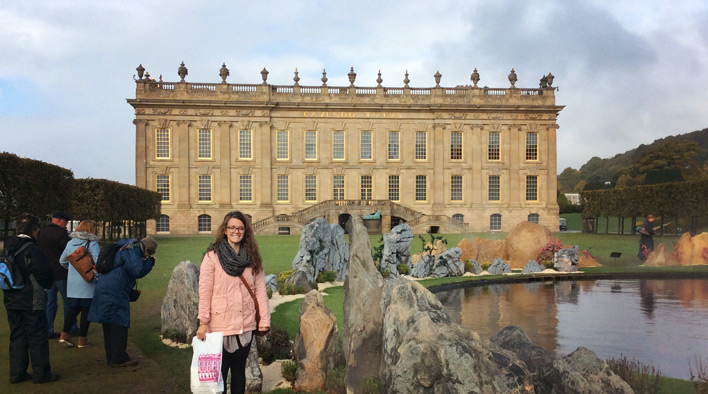 Bex standing in front of Chatsworth House in England. Chatsworth House is a large yellowish building with a grand staircase and a pond. Bex is wearing a chunky black scarf and a thigh-length pink jacket.