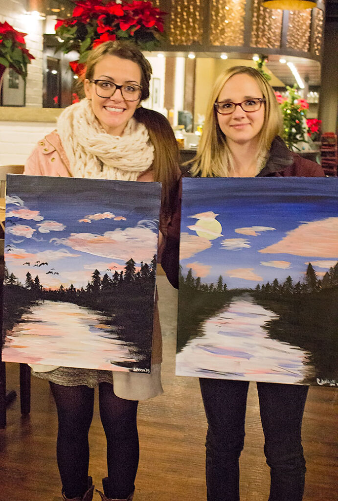 Bex and Chelsey holding their completed paintings. Bex is wearing glasses, a beige knitted scarf, and has her hair in a low ponytail. Chelsey is also wearing glasses, and has blond hair.
