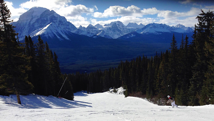 Bex snowboarding down a slope at Lake Louise, surrounded by trees, with the Rocky Mountains in the background. 
