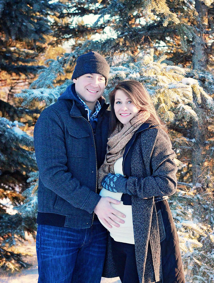 A man with a beanie and blue winter coat standing beside his wife, with his hand on her pregnant belly.