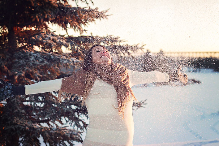 A pregnant woman with a huge smile on her face throwing snow up in the air, creating a glittery effect on the portrait.