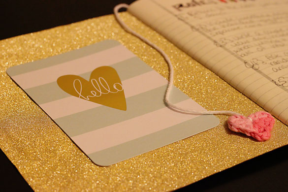 The inside cover of our traveling notebook to keep in touch with friends. It features a gold glitter background, a striped card with a gold heart that says hello, and a crocheted heart as a page marker.