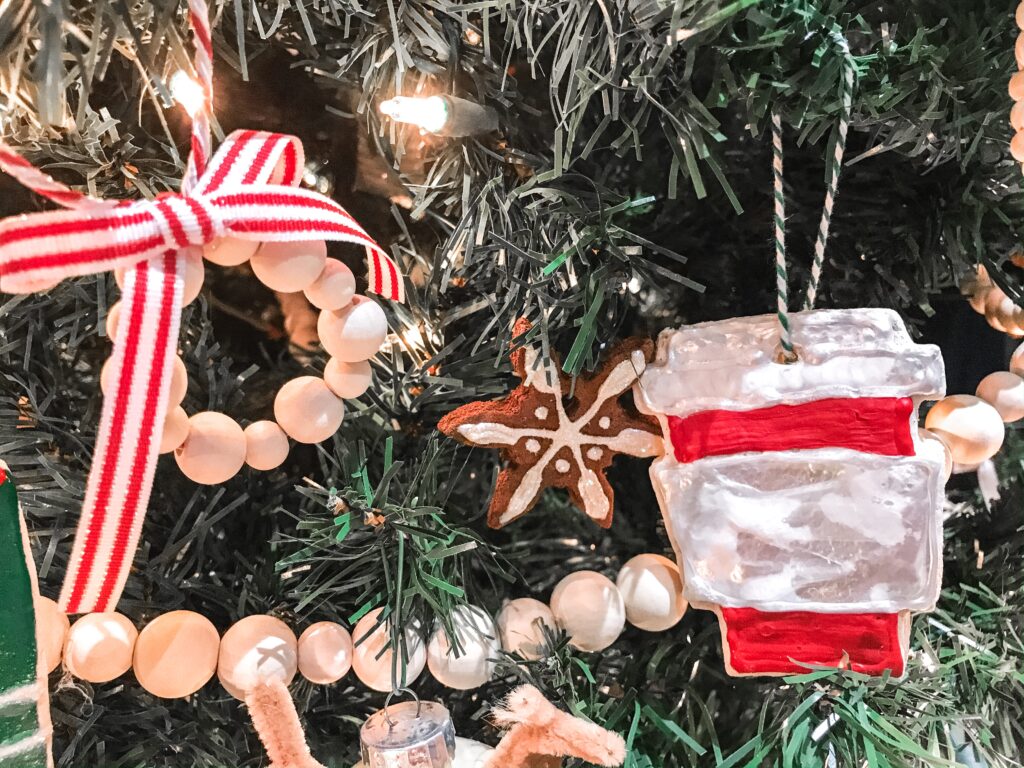 A close up of handmade christmas tree ornaments, one is a circle of wooden beads with a red and white striped ribbon bow and a salt-dough ornament that looks like a red starbucks cup with a white lid.