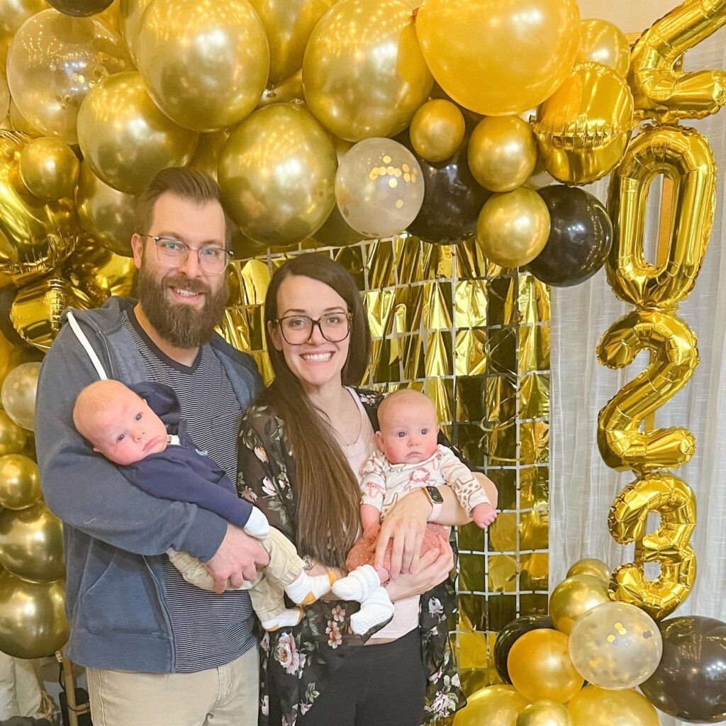 A family of four at a new years party for 2023, standing in front of gold and black balloons with a gold 2023 balloon.