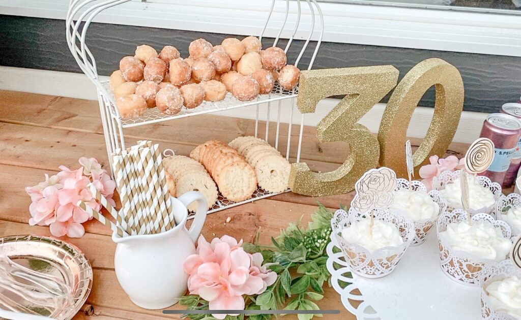 a delicious dessert spread for a creative 30th birthday idea, featuring a cohesive colour-scheme of snacks including cupcakes, donuts, and crackers. Complete with a gold '30' decor piece.