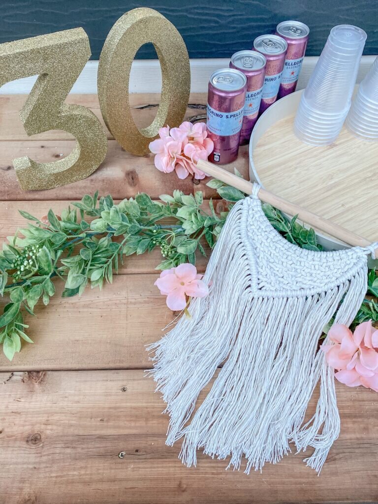 Flatlay of a finished macrame hanging against some greenery, with a gold '30' in the background. 