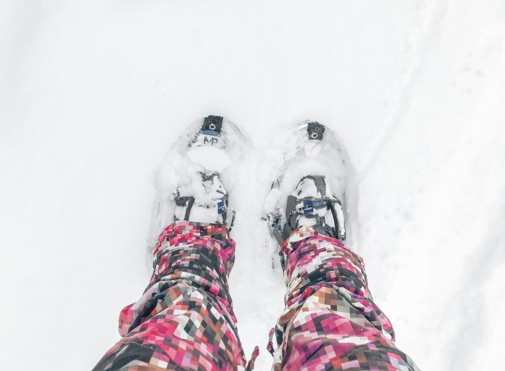 An overhead photo of snowshoes on bex's feet. She is wearing bright pink snowpants with a geometric cube pattern. 